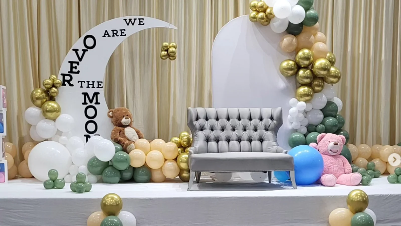 A baby celebration setup by Gubaarewala: a crescent moon sign, lettered balloon boxes and a sage, cream and gold balloon garland arranged across a draped stage